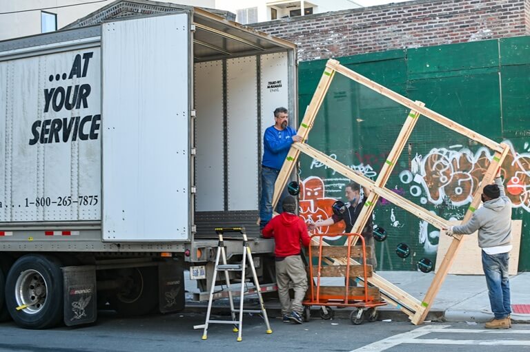 Four movers carrying a large glass window into a white moving truck.